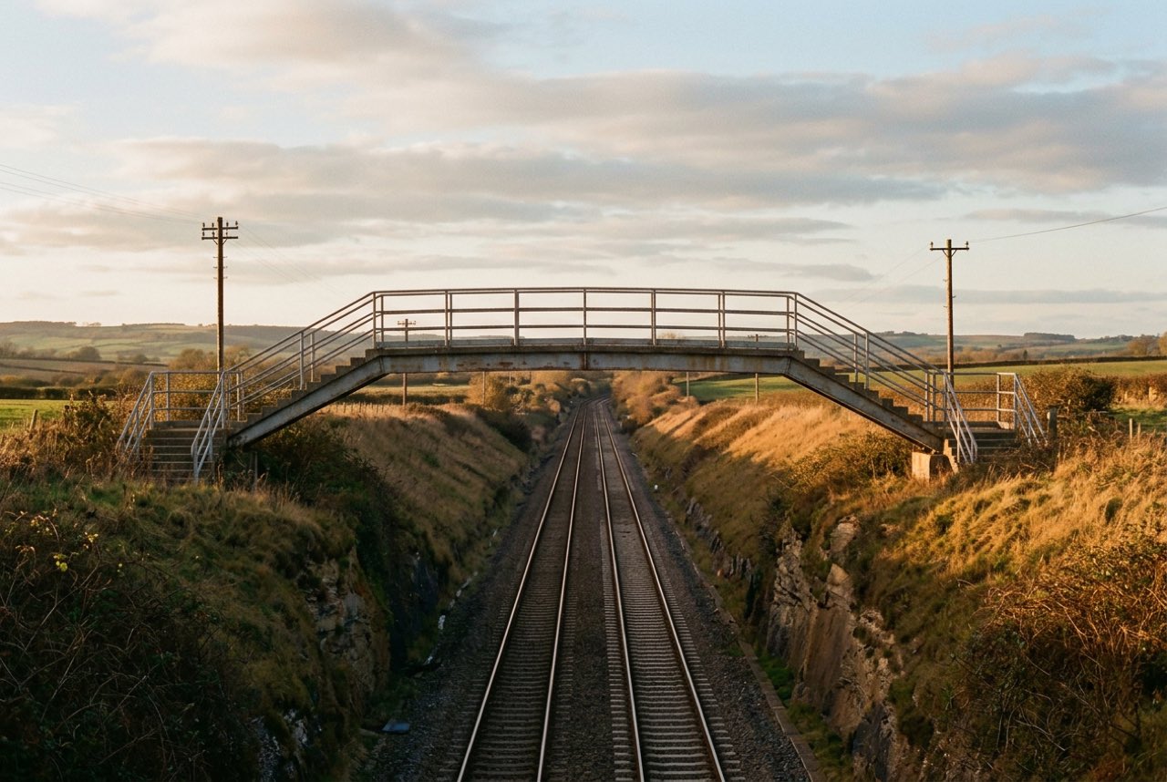 Five in the evening: a footbridge over a railway cutting with the line running away between grass banks, warm low light, no trains