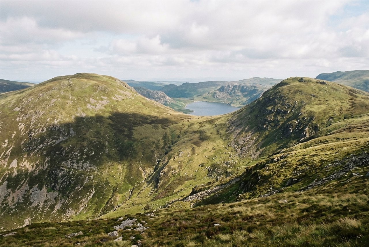 Four in the afternoon: a saddle between two rounded green summits with a lake far below, cloud shadow moving across the slope